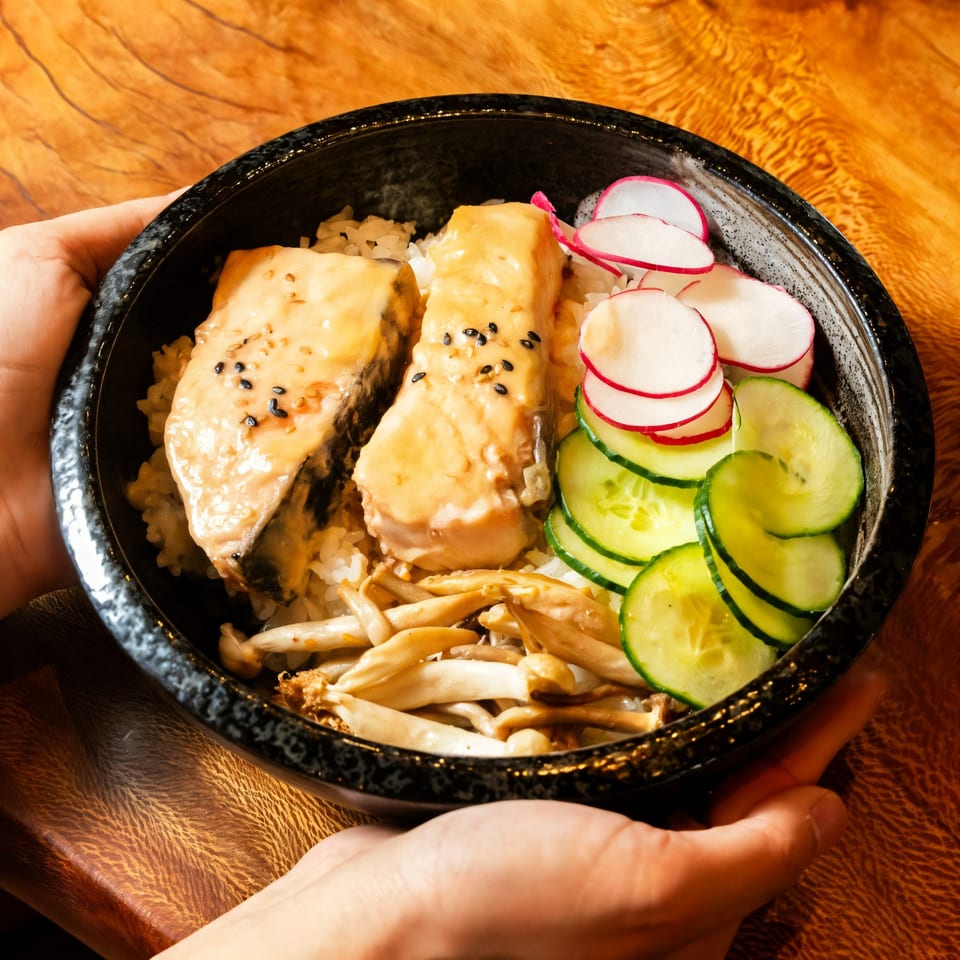 Hands holding a black bowl of Kakui Sushi's salmon, rice, mushrooms, cucumbers, and radishes.