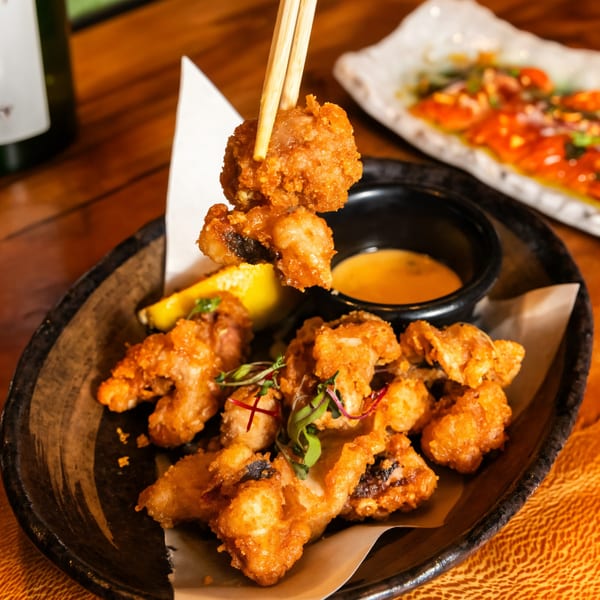 Chopsticks holding a piece of crispy fried chicken over a plate of more chicken, sauce, and lemon.